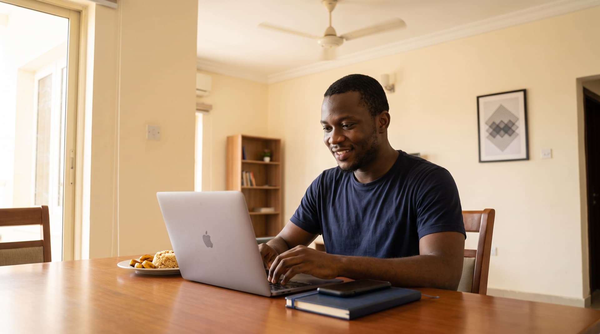 Nigerian freelancer typing on a laptop at a dining table in a Lagos apartment